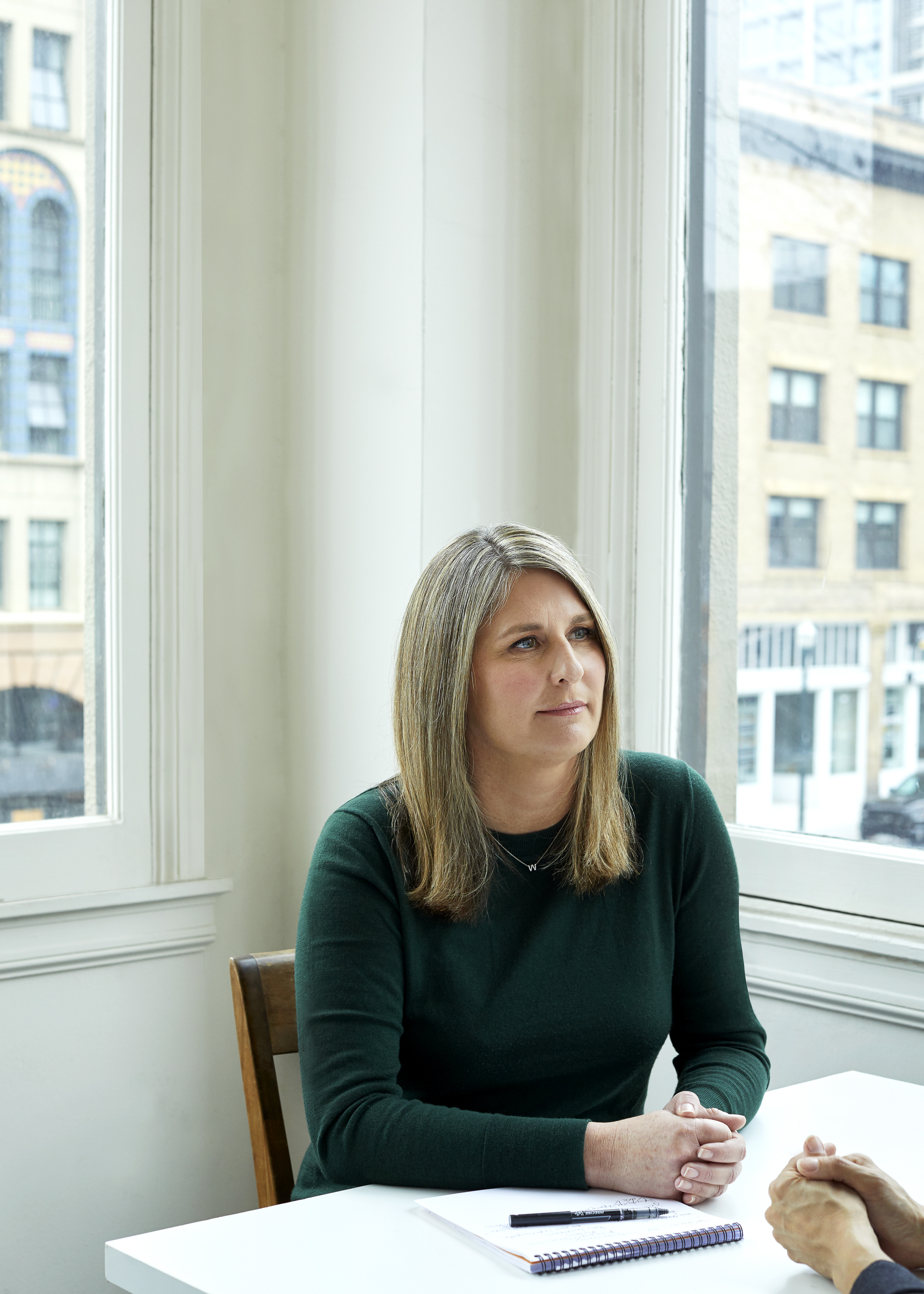 Aspen Baker seated at desk. Behind her are two windows looking out onto buildings in the distance.
